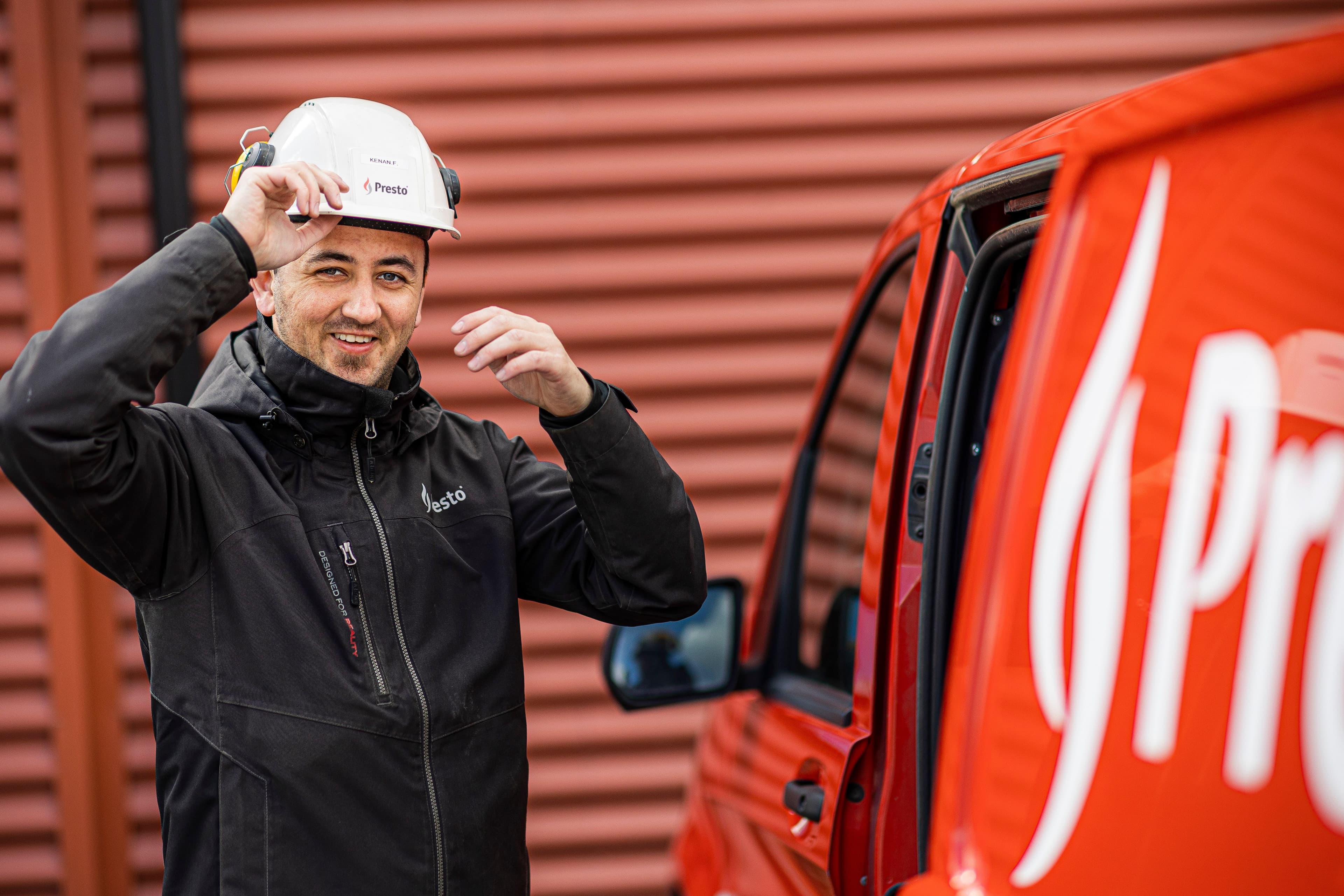 Person i arbetskläder som justerar sin skyddshjälm bredvid en röd Presto‑servicebil