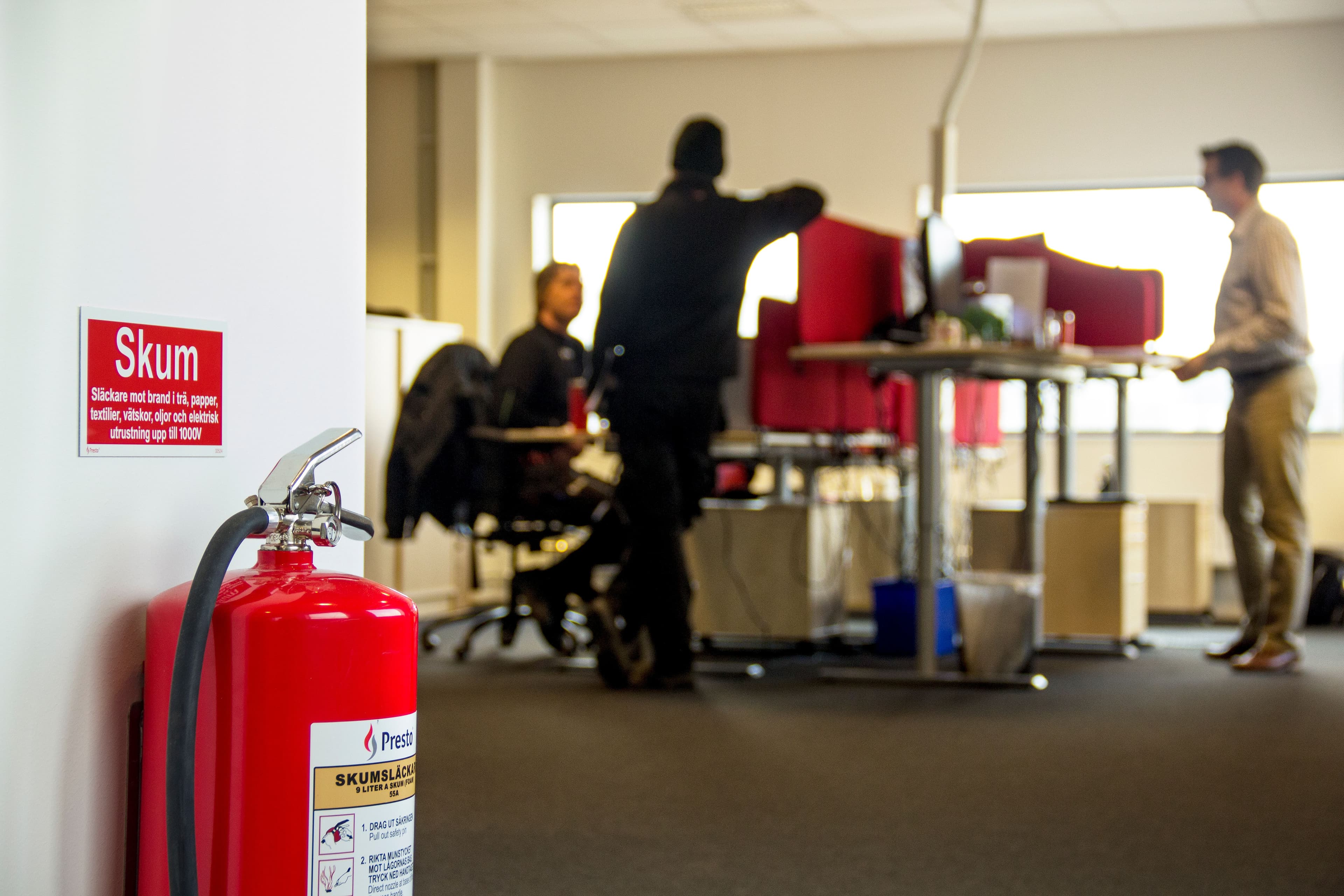 Brandsläckare med skummärkning i förgrunden på ett kontor där flera personer arbetar vid skrivbord i bakgrunden